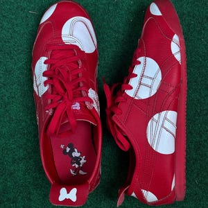 Puede incluir: Par de zapatillas rojas con lunares blancos. Los zapatos tienen cordones y suelas rojas. El interior presenta un gr&aacute;fico de Minnie Mouse. Los zapatos se exhiben sobre una superficie verde.