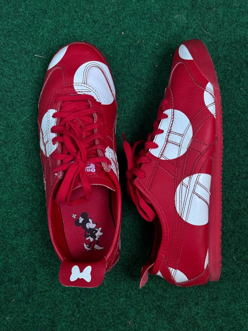 Puede incluir: Par de zapatillas rojas con lunares blancos. Los zapatos tienen cordones y suelas rojas. El interior presenta un gr&aacute;fico de Minnie Mouse. Los zapatos se exhiben sobre una superficie verde.