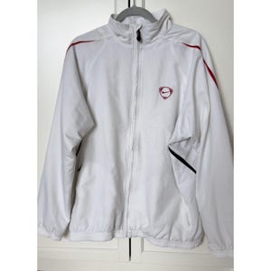 May include: White Nike track jacket with a full zipper, red accents on the shoulders, and a red and white Nike logo on the chest. The jacket has a high collar and black accents on the sleeves.