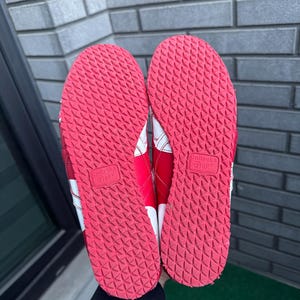 Puede incluir: Par de zapatillas deportivas rojas y blancas, vistas desde las suelas. Las suelas son de goma roja texturizada con un patr&oacute;n repetitivo. Rayas blancas y el nombre de la marca son visibles en los laterales.