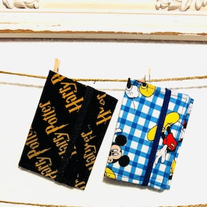May include: Two small fabric notebooks hanging from a clothesline. One is black with gold "Harry Potter" text. The other is blue and white checkered with a Mickey Mouse design and a blue elastic band.