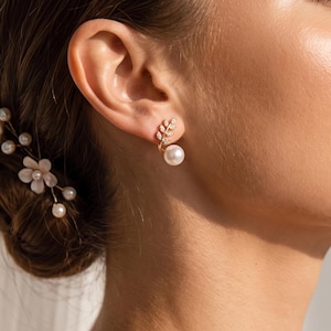 Pode incluir: Um brinco dourado com um design em forma de folha incrustado com pequenas pedras transparentes e uma pérola branca pendente. O brinco é usado no lóbulo da orelha. Um acessório de cabelo a condizer com pequenas pérolas brancas é visível.
