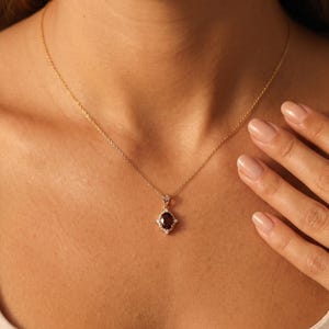 Può includere: Una collana a catena dorata con un ciondolo in pietra preziosa rosso scuro di forma ovale. Il ciondolo è circondato da piccole pietre chiare e scintillanti incastonate in una montatura dorata, creando un aspetto delicato e raffinato.