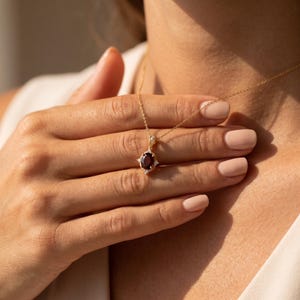 May include: A gold chain necklace featuring an oval-shaped dark red gemstone pendant. The pendant is accented with small, clear, sparkling stones set in a decorative gold frame. The necklace rests against the skin, showcasing the delicate metal link chain.