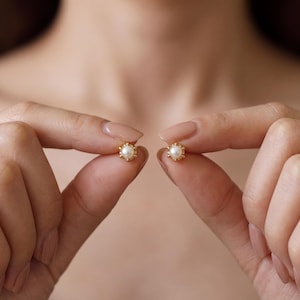 Puede incluir: Un par de pendientes de botón en oro, cada uno con una perla blanca central rodeada por un delicado marco dorado con cuentas. Los pendientes son pequeños, circulares y muestran un acabado metálico pulido de aspecto refinado.