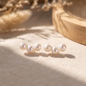 May include: A pair of ear climber earrings, each featuring three round, lustrous white pearls arranged in a curved, ascending line. The pearls are set on a thin metal base, creating a delicate and structured design that follows the earlobe contour.