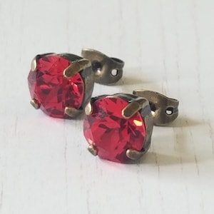 May include: A pair of stud earrings featuring large, faceted, round, ruby-red gemstones. The stones are set in antique bronze-colored metal settings. The earring posts are also bronze-colored. The earrings are displayed on a white, textured surface.