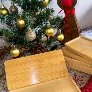 May include: A set of four natural bamboo trays, stacked on top of each other. The trays are rectangular and have a smooth, polished finish. The trays are in front of a Christmas tree decorated with gold ornaments and a wooden reindeer figurine.