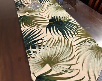 Puede incluir: Un camino de mesa beige con un patrón de hojas tropicales en tonos verdes. El camino de mesa está sobre una mesa de madera con una tapa de vidrio.
