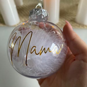 Könnte beinhalten: Ein transparenter Weihnachtsbaumschmuck aus Glas mit einer goldenen Aufschrift "Mama" und weißen Federn im Inneren.