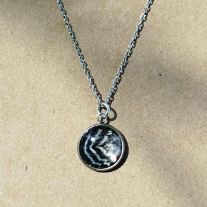Puede incluir: Un collar de cadena de metal plateado con un colgante circular que presenta un patrón de chevrón en blanco y negro. El collar se muestra sobre una tarjeta rectangular de color canela con una pequeña silueta de pájaro negro impresa en el centro inferior.
