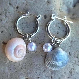 Könnte beinhalten: Ein Paar silberne Creolen mit hängenden Anhängern. Ein Ohrring hat ein glattes, blassrosa spiralförmiges Muschelgehäuse und eine kleine weiße Perle. Der andere Ohrring zeigt eine strukturierte, graue fächerförmige Muschel und eine kleine weiße Perle.