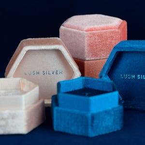 May include: A collection of hexagonal velvet ring boxes in various colors: light pink, peach, and blue. Each box has a contrasting interior and the text "LUSH SILVER" printed inside. The boxes are arranged on a dark blue surface.