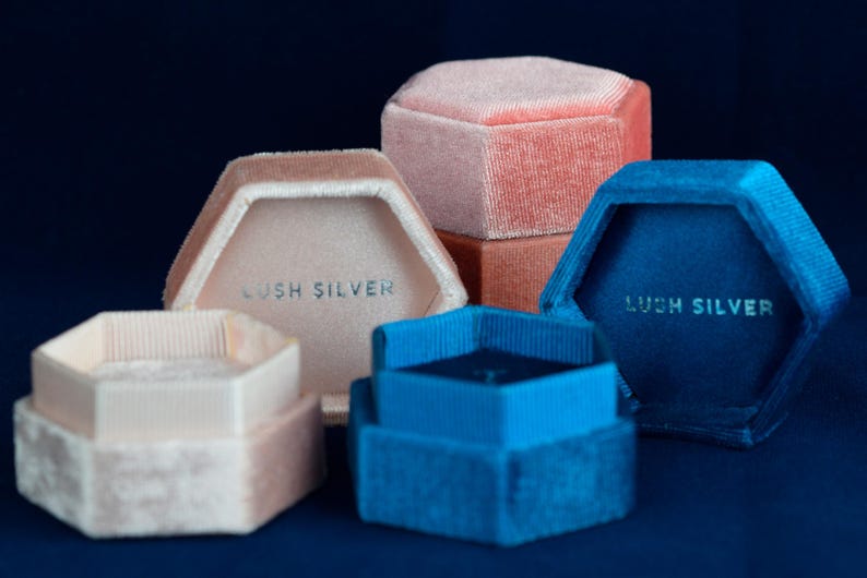 May include: A collection of hexagonal velvet ring boxes in various colors: light pink, peach, and blue. Each box has a contrasting interior and the text "LUSH SILVER" printed inside. The boxes are arranged on a dark blue surface.