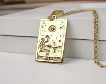 May include: Gold-colored pendant necklace featuring a rectangular tarot card design. The card depicts a woman pouring water, with the words "THE STAR" at the bottom. The pendant hangs from a delicate gold chain.