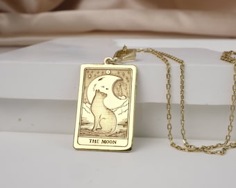 May include: Gold necklace with a rectangular pendant featuring a tarot card design. The card depicts a cat sitting under a crescent moon with stars in the background. The card reads "THE MOON".