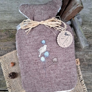 Könnte beinhalten: Eine braune Filz-Wärmflasche mit einem weißen flauschigen Futter. Die Flasche hat ein kleines gesticktes Design eines Vogels, der auf einem Zweig mit blauen Blumen sitzt. Ein braunes Etikett mit weißem Text lautet "zauberhaft!  Handgefertigte  Schönes  für Dich".