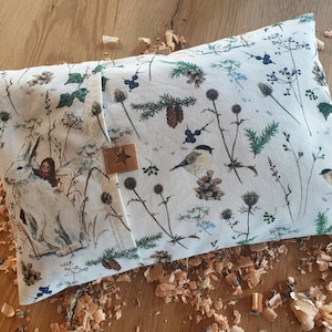 Könnte beinhalten: Rechteckiges Kissen mit einem skurrilen Winterwald-Druck. Das Design zeigt einen weißen Hasen mit einer Fee, Vögeln, Tannenzapfen und verschiedenen Pflanzen in Grün-, Blau- und Brauntönen. Ein braunes Lederetikett mit einem Stern ist angebracht.