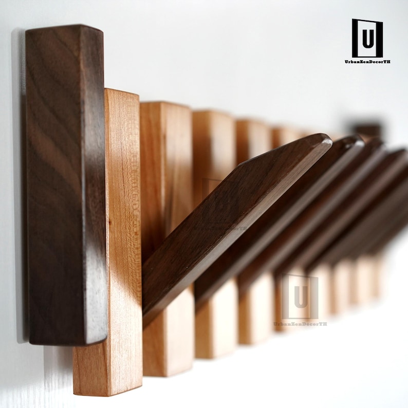 May include: A wooden wall-mounted coat rack with a modern design. The rack features a series of rectangular pegs that can be flipped up or down. The pegs are made of dark brown and light brown wood. The rack is mounted on a white wall.