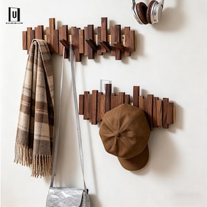 Op de afbeelding: Twee houten kapstokken met een uniek ontwerp, met verticale houten blokken. Eén kapstok houdt een bruine sjaal en een zilveren tas, de andere een bruine pet. Een koptelefoon hangt erboven. De kapstokken zijn aan een witte muur bevestigd.