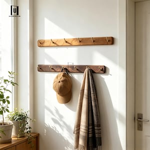 May include: Two wooden coat racks with brass hooks mounted on a white wall. The top rack is a lighter wood, while the bottom rack is a darker wood. A brown baseball cap and a tan and brown scarf hang from the bottom rack.