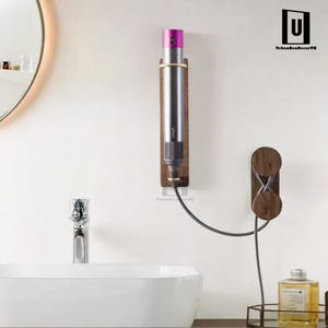 Op de afbeelding: Een houten wandbevestiging voor een föhn met een snoerorganizer. De föhn is donkergrijs met een roze accent. De beugel is gemaakt van donker hout en heeft een gouden accent.