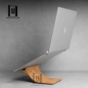 May include: A wooden laptop stand with a curved design, supporting a silver laptop. The stand is made of natural wood and has a smooth finish. The laptop is closed and has a silver Apple logo on the back.