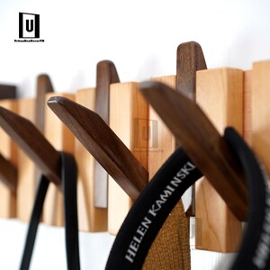 May include: Wooden wall-mounted coat rack with a natural wood finish and dark brown hooks. The rack features a series of rectangular blocks with angled hooks, designed for hanging coats, hats, or bags. The design is modern and minimalist.