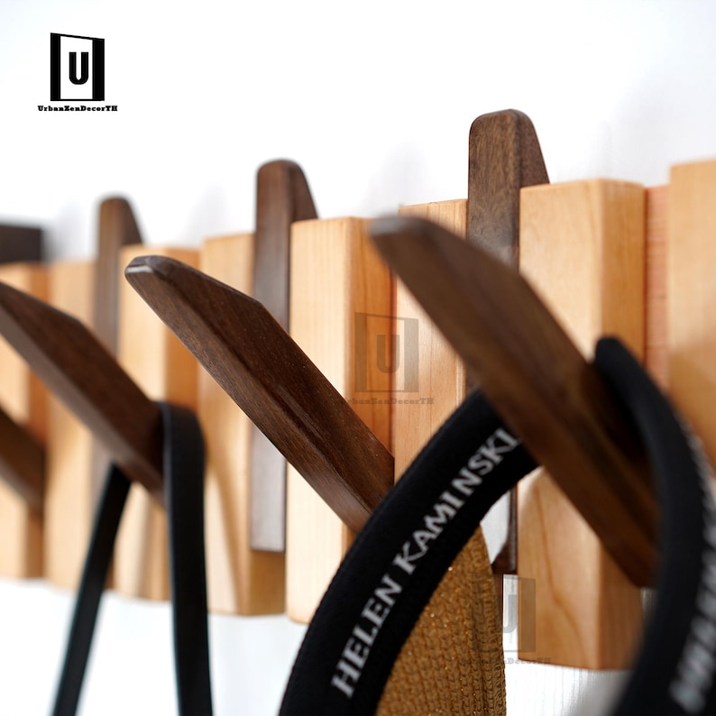 May include: Wooden wall-mounted coat rack with a natural wood finish and dark brown hooks. The rack features a series of rectangular blocks with angled hooks, designed for hanging coats, hats, or bags. The design is modern and minimalist.