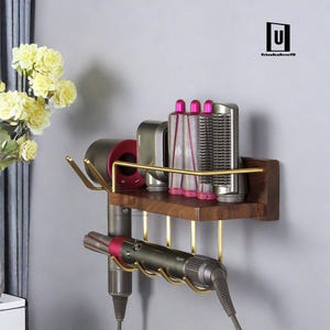 May include: A wooden wall-mounted shelf with a gold metal rack and hooks for storing hair styling tools. The shelf has a hairdryer, curling iron, and attachments on it.