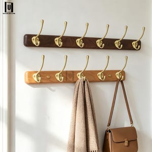 Könnte beinhalten: Zwei horizontale Wandgarderoben mit Haken aus poliertem Messing. Die obere Leiste besteht aus dunklem Walnussholz, die untere aus hellem Eichenholz. Ein beigefarbener, strukturierter Schal und eine braune Ledertasche hängen an den Haken.
