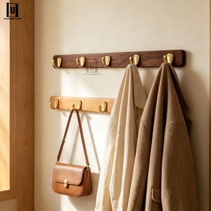 Peut inclure: Deux porte-manteaux muraux horizontaux en bois avec des crochets en métal doré. L'étagère supérieure est en bois de noyer foncé avec cinq crochets, et l'étagère inférieure est en bois de chêne clair avec trois crochets. Un sac à main en cuir beige et deux manteaux sont suspendus aux crochets.