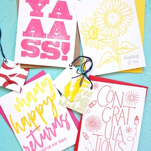 May include: A collection of greeting cards with various designs and text. One card features the word "YAASS!" in pink, another has a yellow sunflower, and a third says "Many happy returns". A fourth card says "CONGRATULATIONS".