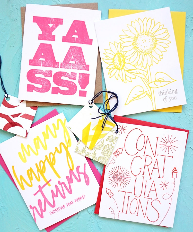May include: A collection of greeting cards with various designs and text. One card features the word "YAASS!" in pink, another has a yellow sunflower, and a third says "Many happy returns". A fourth card says "CONGRATULATIONS".