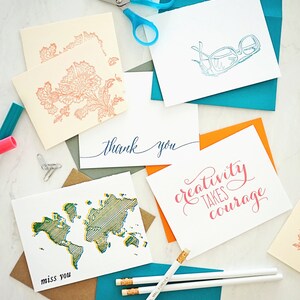 May include: A collection of greeting cards with various designs and messages. Cards feature floral patterns, glasses, a world map, and the phrases "thank you", "miss you", and "creativity takes courage". Pencils and scissors are also present.