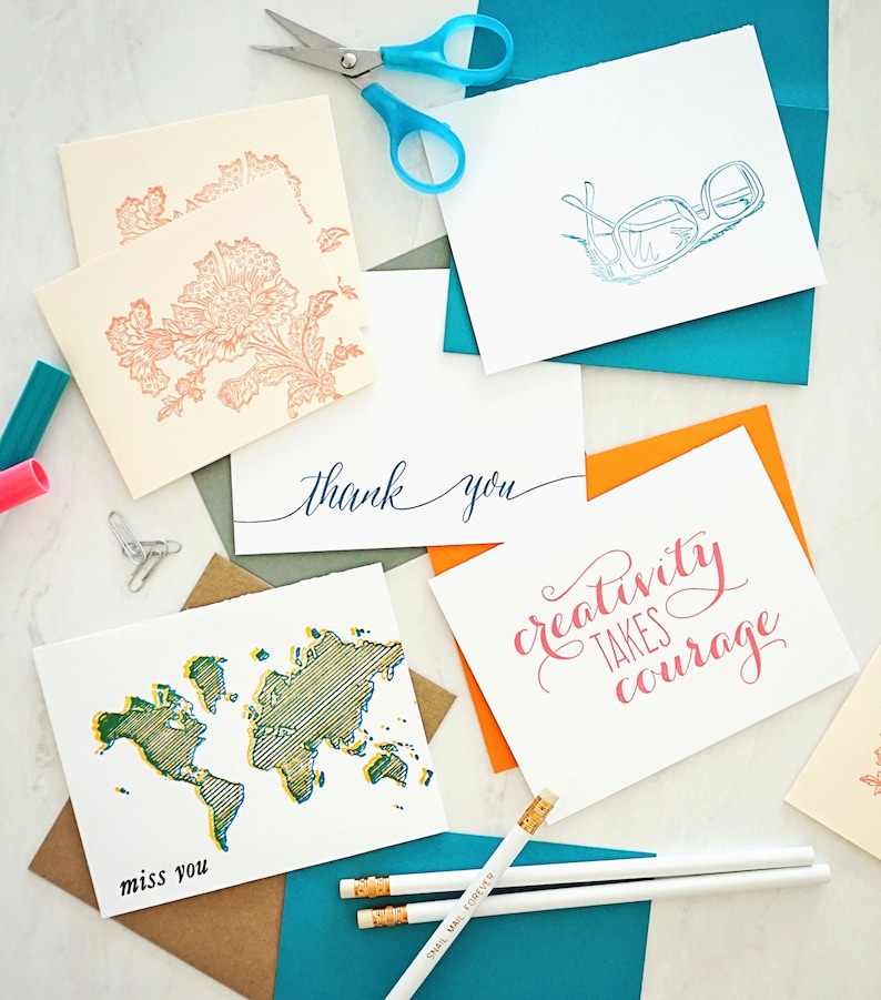 May include: A collection of greeting cards with various designs and messages. Cards feature floral patterns, glasses, a world map, and the phrases "thank you", "miss you", and "creativity takes courage". Pencils and scissors are also present.