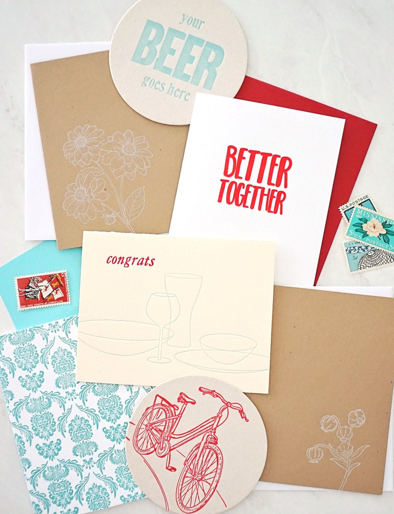 May include: Assortment of greeting cards and coasters. One coaster reads "your BEER goes here" in blue. Other cards feature the text "BETTER TOGETHER" and "congrats". Floral and bicycle designs are also present.