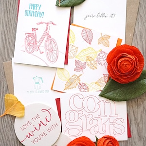May include: A collection of greeting cards with various designs and text, including "Happy Birthday!" and "Congrats." The cards feature illustrations of a bicycle, leaves, and a coffee maker, along with decorative paper flowers and coasters.