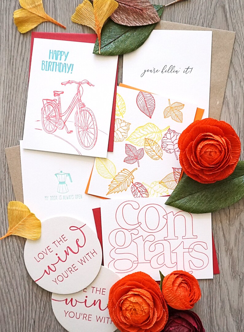 May include: A collection of greeting cards with various designs and text, including "Happy Birthday!" and "Congrats." The cards feature illustrations of a bicycle, leaves, and a coffee maker, along with decorative paper flowers and coasters.