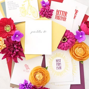 May include: Greeting cards with various designs and text, including "Better Together," "Best Mom Ever," and a bicycle illustration. The cards are surrounded by colorful flowers and envelopes.