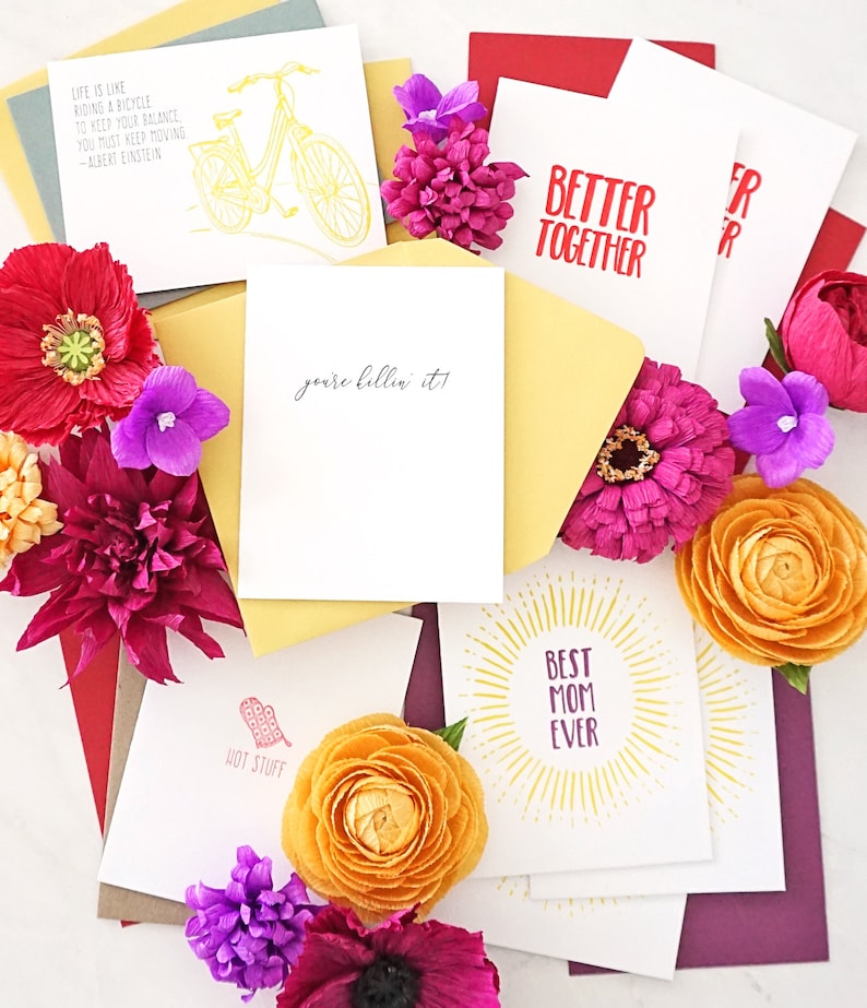 May include: Greeting cards with various designs and text, including "Better Together," "Best Mom Ever," and a bicycle illustration. The cards are surrounded by colorful flowers and envelopes.
