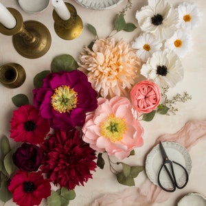 May include: A collection of paper flowers in various colours and styles, including pink, red, white, and peach. The flowers are arranged in a bouquet-like formation, with some flowers overlapping others. The flowers are made of paper and have a delicate, handmade appearance.