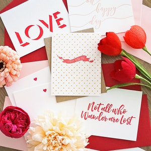 May include: A collection of greeting cards and envelopes in shades of red, pink, and white. Some cards feature text like "LOVE", "oh happy day", and "Not all who wander are lost". Decorative paper flowers and red tulips are also present.