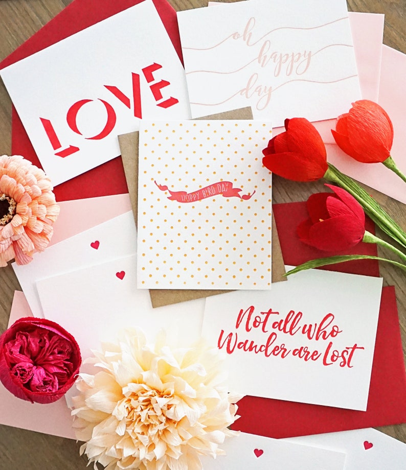 May include: A collection of greeting cards and envelopes in shades of red, pink, and white. Some cards feature text like "LOVE", "oh happy day", and "Not all who wander are lost". Decorative paper flowers and red tulips are also present.