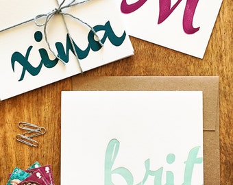 May include: Three white cards with handwritten names in colorful paint. The names are "Xina", "Brit", and "M". The cards are on a wooden surface with a brown envelope and some stamps.