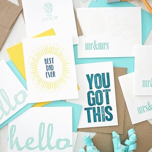 May include: A collection of greeting cards with various designs and messages. The cards feature phrases like "hello", "Best Dad Ever", and "You Got This". The color palette includes white, teal, yellow, and brown.