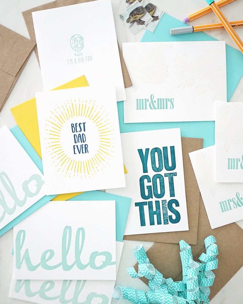 May include: A collection of greeting cards with various designs and messages. The cards feature phrases like "hello", "Best Dad Ever", and "You Got This". The color palette includes white, teal, yellow, and brown.