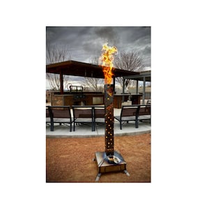 May include: A tall, metal fire pit with a decorative design and a flame burning inside. The fire pit is standing on a patio with chairs and a covered outdoor kitchen in the background.