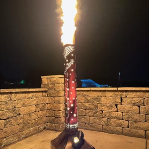 May include: A tall, metal fire pit with a flame shooting upwards. The fire pit has a red and black design with star cutouts and the words "We the People". The base has the text "GRUNT FABWORKS".