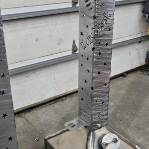 May include: A metal fire pit with a cutout design of the American flag and the words "We the People". The fire pit is made by GRUNT FAB and has a square base.
